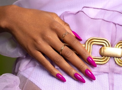 Mão com unhas rosa vibrante, anéis dourados e roupa lilás com cinto dourado