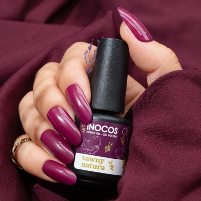 Frasco de verniz gel Inocos na cor vinho com mão a segurar unhas compridas na mesma cor e fundo de tecido vinho.