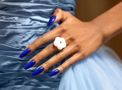 Mão com unhas azuis e anel de flor branco sobre vestido azul