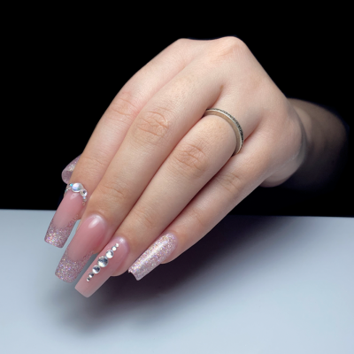Mão feminina com unhas postiças rosadas com glitter e cristais, e anel prateado simples.
