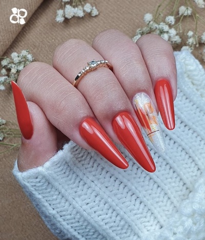 Mão feminina com unhas vermelhas compridas e decoração floral numa unha, com anel dourado e roupa de malha branca.