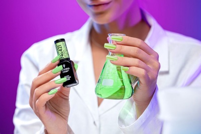 Pessoa com unhas verdes segurando frasco de verniz Victoria Vynn e frasco de laboratório com líquido verde