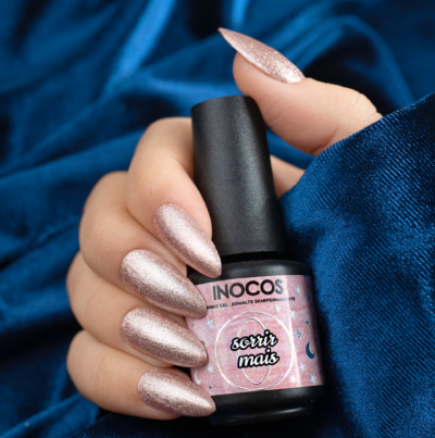 Mão com unhas compridas pintadas segurando frasco de verniz Inocos 'sorrir mais' sobre fundo azul.