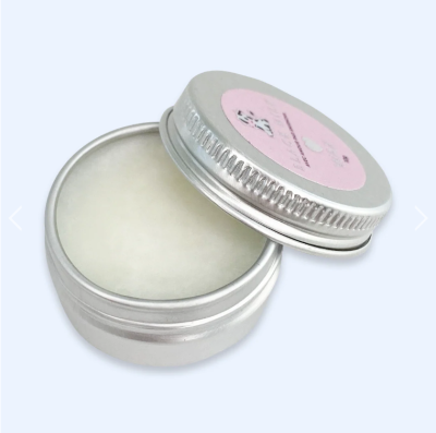 Pote metálico prateado aberto com pomada branca e tampa com etiqueta rosa.