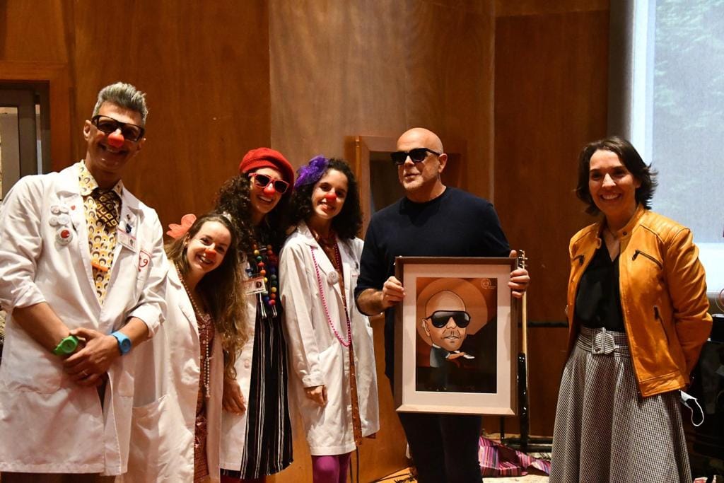 Seis pessoas em interior com roupas coloridas e quadro de pessoa calva com óculos escuros