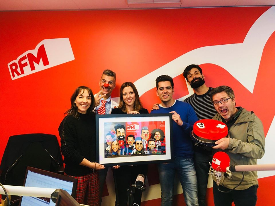 Grupo de seis pessoas num estúdio de rádio com logo RFM, segurando quadro e objeto vermelho com rosto desenhado