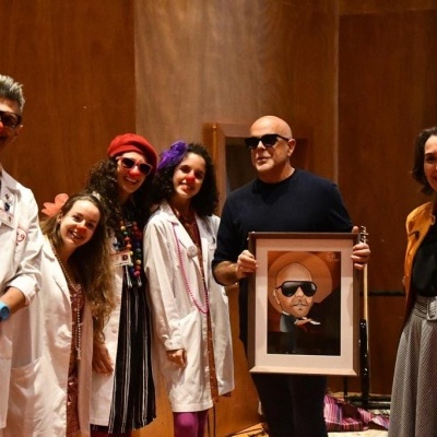 Seis pessoas em interior com roupas coloridas e quadro de pessoa calva com óculos escuros