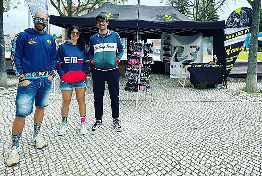 Três pessoas com roupa casual em frente a tendas pretas com equipamentos esportivos e banners com texto EM3 e M3.