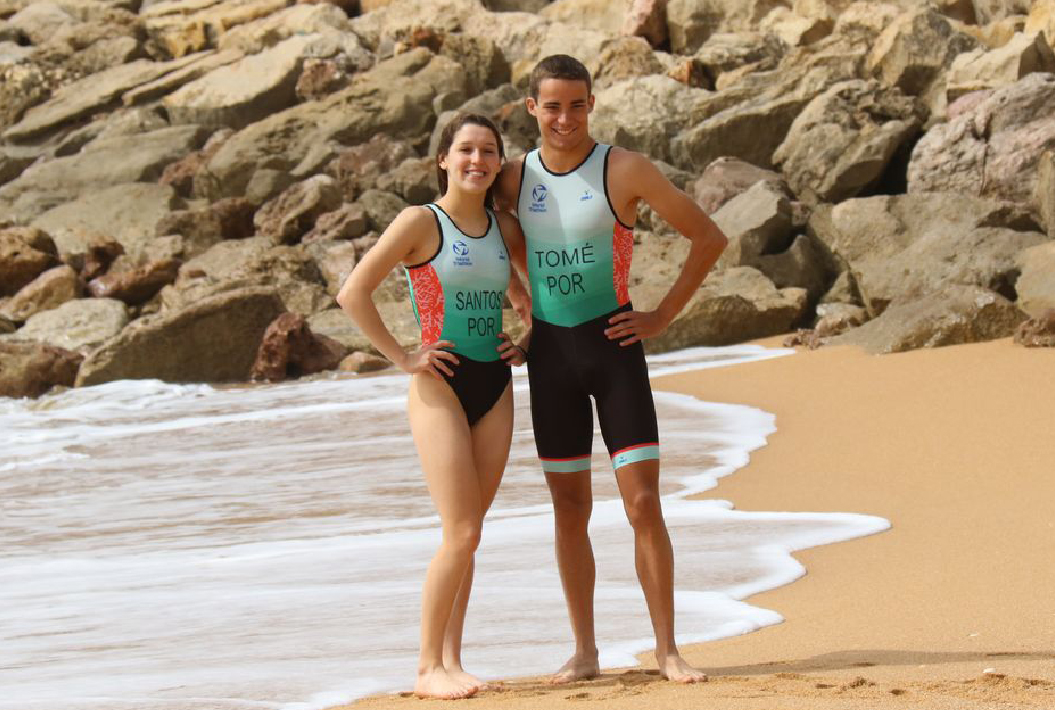 EM3 veste Federação de Triatlo de Portugal Dois jovens em fatos de triatlo pretos com inscrições verdes e vermelhas na praia