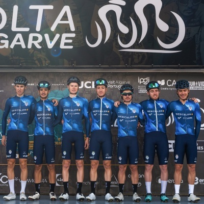 Equipa de ciclismo com fatos azuis e pretos com logos, capacetes, posando em frente a painel da Volta ao Algarve.