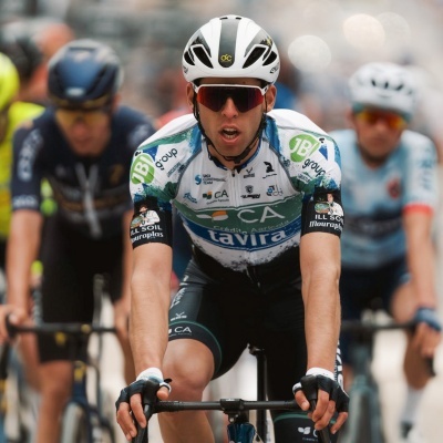 Ciclista com capacete branco e roupas técnicas brancas, verdes e pretas com logotipos, a pedalar em competição