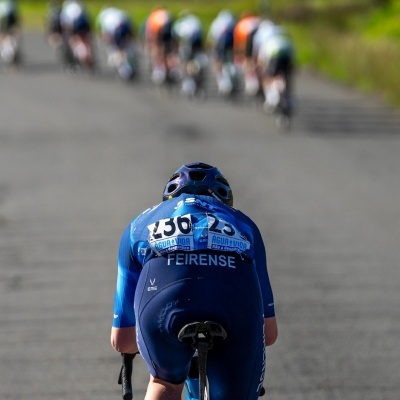 Ciclista com equipamento azul e capacete a pedalar em estrada com outros ciclistas ao fundo