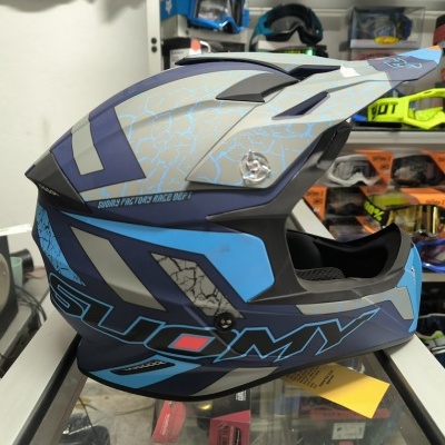 Capacete de motocross azul com texto SUOMY numa loja com óculos de motocross ao fundo
