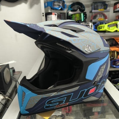 Capacete integral azul, cinzento e preto com texto SUOMY SAFETY junto a vários óculos de proteção numa loja