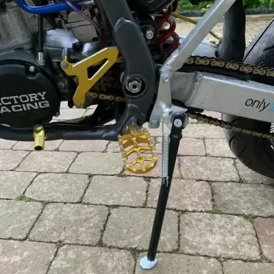 Close-up do motor e transmissão de uma motocicleta com pedal dourado e apoio lateral preto