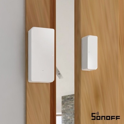 Sensor branco Sonoff numa porta de madeira clara, parede cinza e parede de pedra ao fundo