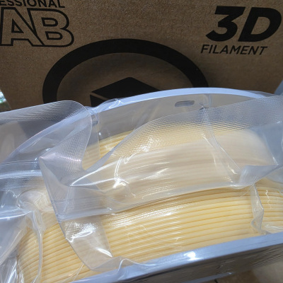 Rolo de filamento 3D cor bege claro embalado em saco plástico transparente com caixa castanha ao fundo
