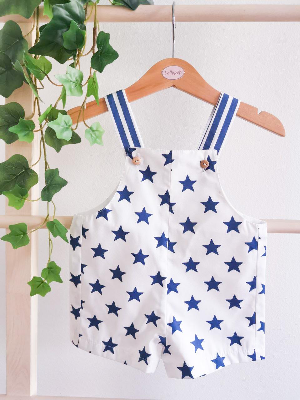Macacão de bebé branco com estrelas azuis e alças azuis com listras brancas pendurado num cabide de madeira