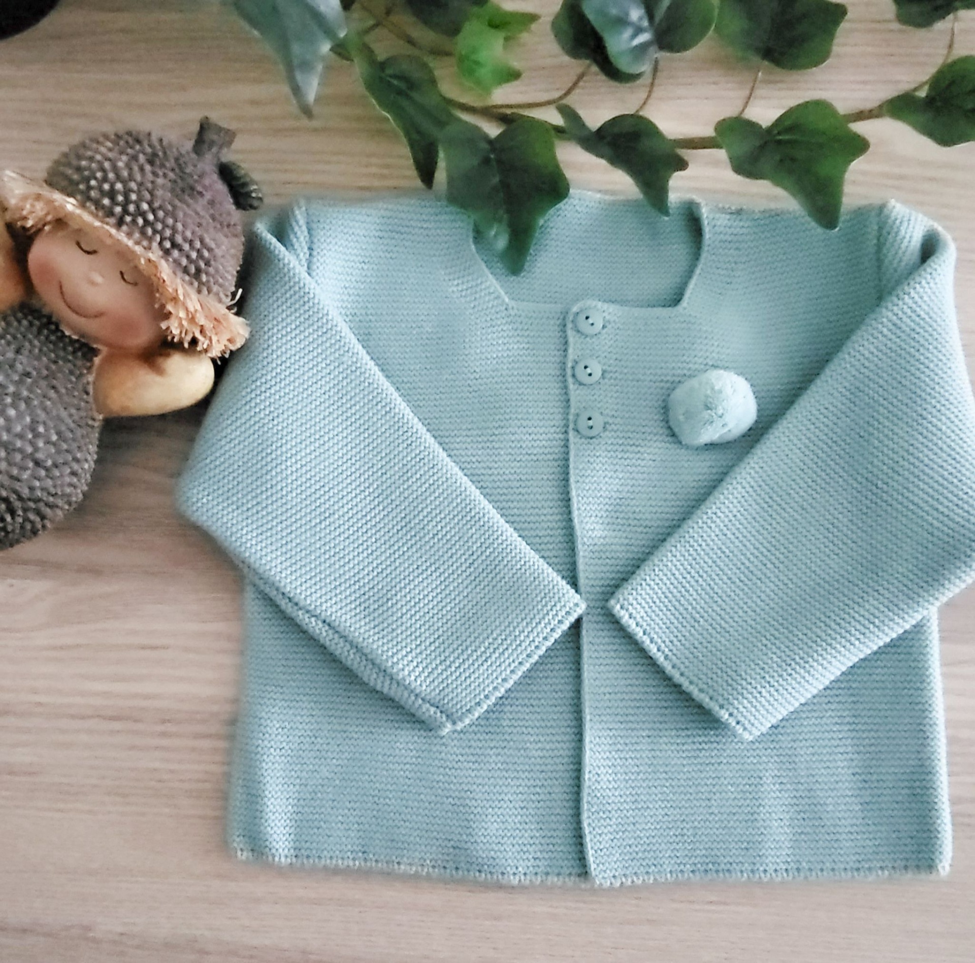 Casaco de malha azul claro com botões e pompom sobre superfície de madeira com boneco e folhas verdes