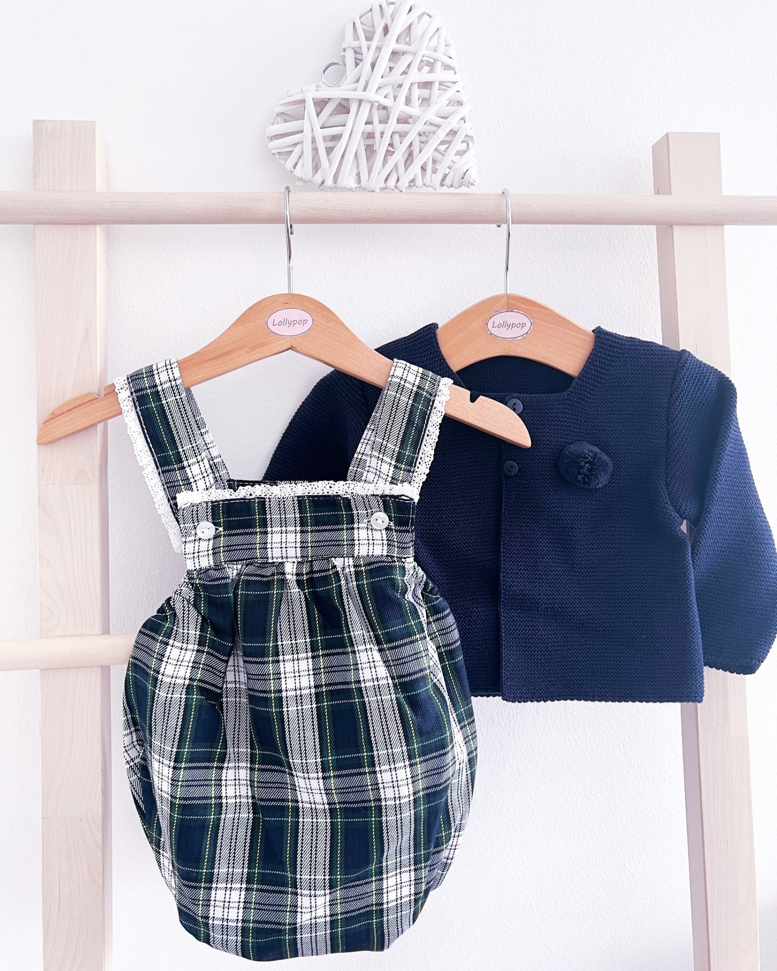 Conjunto de roupa infantil com macacão xadrez verde e casaco azul escuro pendurados em cabides numa estrutura de madeira clara.