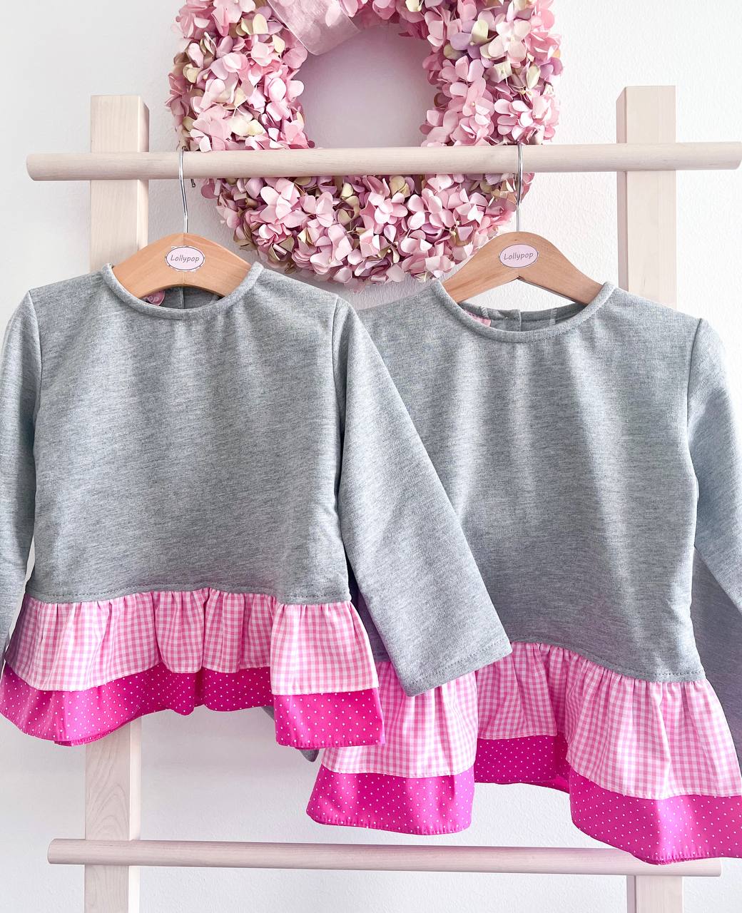 Duas blusas infantis cinza com bainha rosa penduradas em cabides de madeira, com coroa de flores cor-de-rosa ao fundo