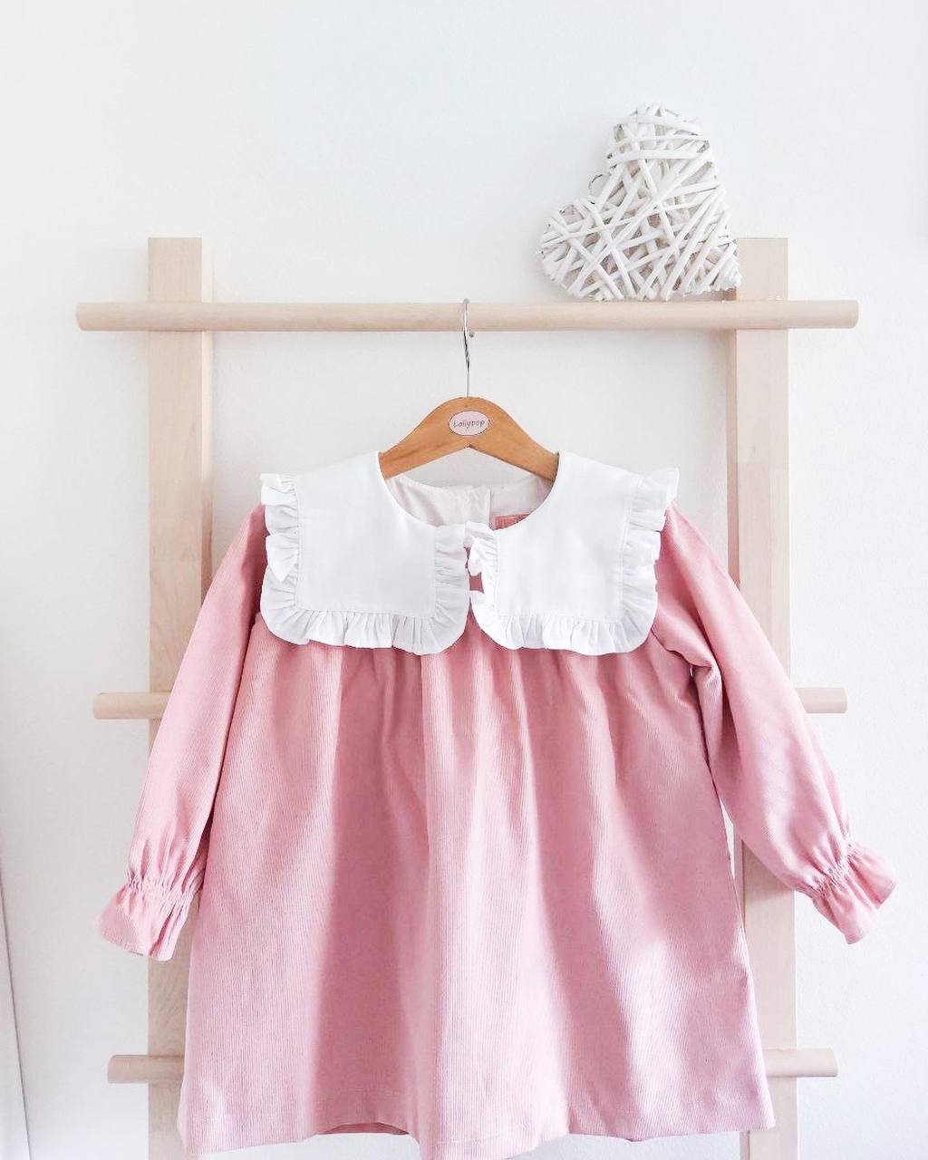 Vestido infantil rosa com gola branca grande e folhos pendurado num cabide de madeira