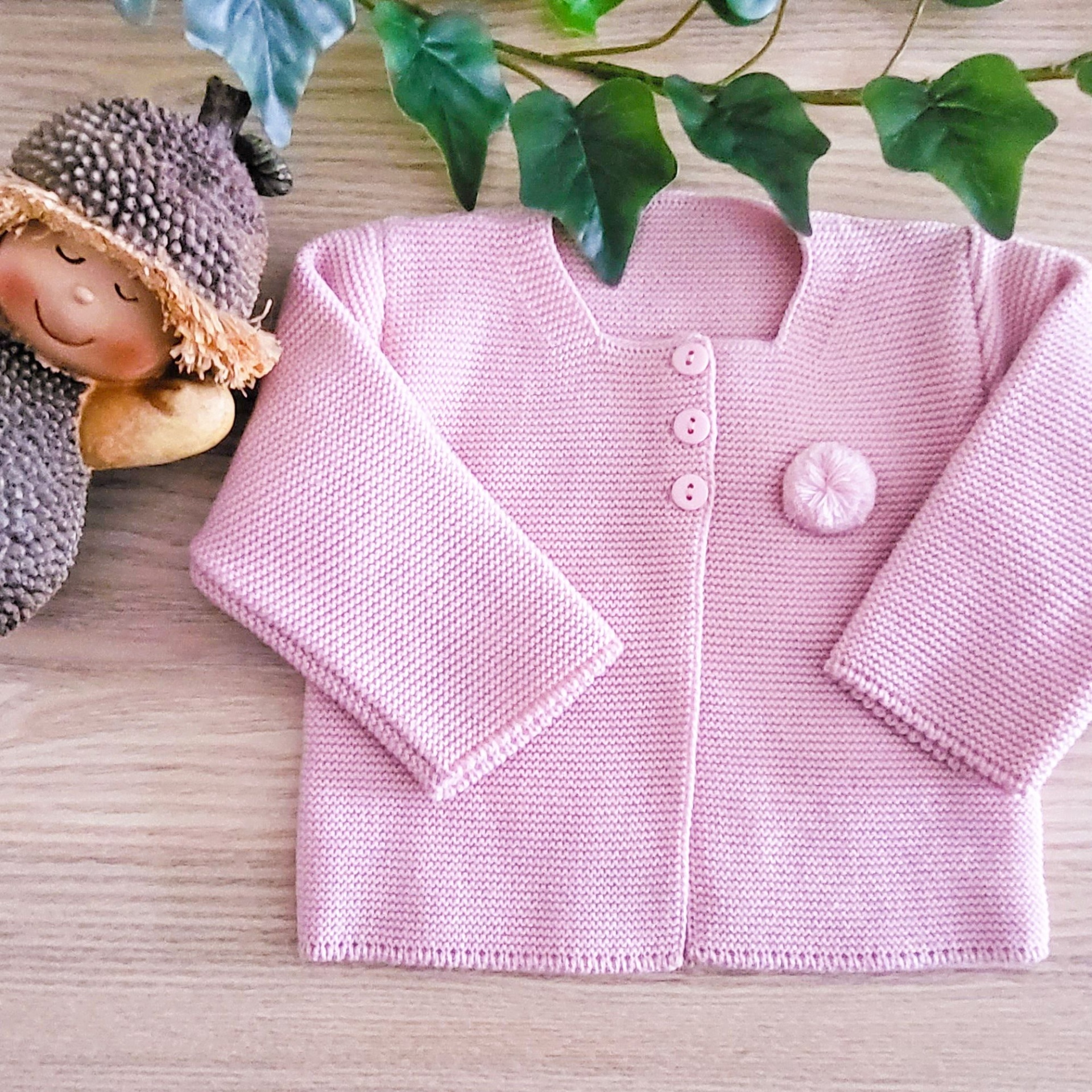 Casaco de malha rosa claro para bebé com botões e aplique de flor, sobre madeira clara