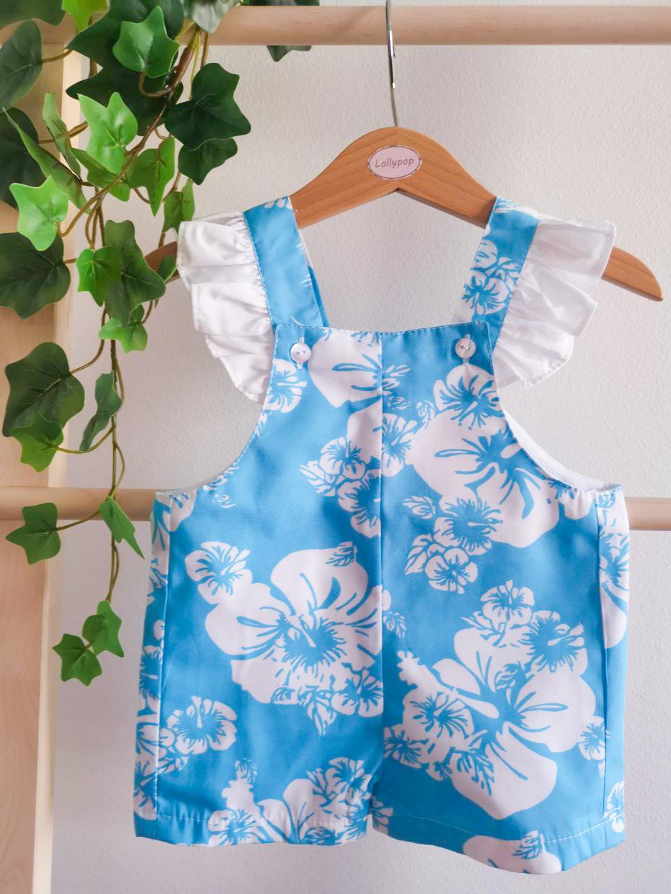 Macacão azul com flores brancas para criança pendurado num cabide com planta ao lado