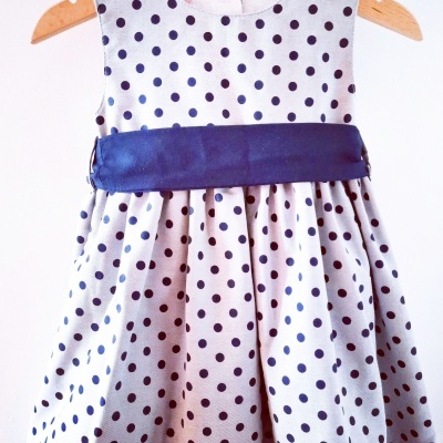 Vestido infantil branco com bolinhas azuis e faixa azul escura pendurado num cabide