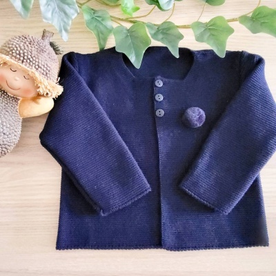 Casaco de malha azul escuro com botões e pompom sobre mesa de madeira, decorado com folhas verdes e figura de criança vestida de castanho