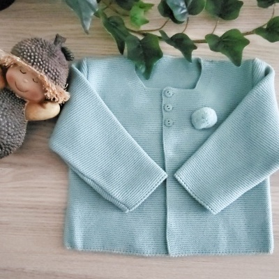 Casaco de malha azul claro com botões e pompom sobre superfície de madeira com boneco e folhas verdes