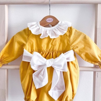 Macacão amarelo para bebé com gola branca e grande laço, pendurado em cabide de madeira.