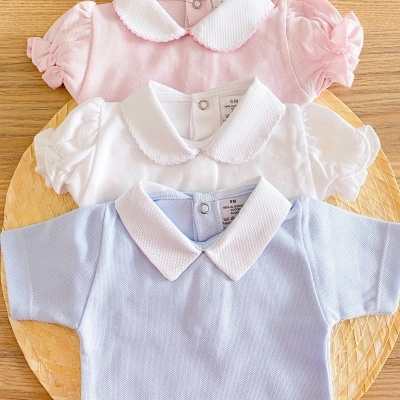 Três bodies de bebé em rosa, branco e azul claro com golas brancas e mangas curtas, sobre base redonda de vime.