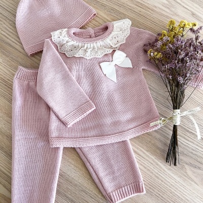 Conjunto de roupa de bebé rosa clara com gola rendada e laço, calças e gorro com nó, ao lado ramo de flores secas