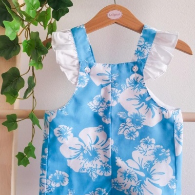 Macacão azul com flores brancas para criança pendurado num cabide com planta ao lado