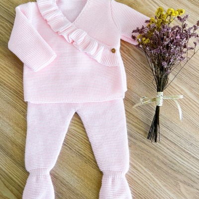 Conjunto de roupa de bebê rosa claro em tricô com folho e botão dourado, calças com pé, e ramo de flores secas ao lado