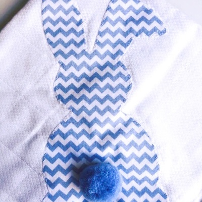 Pano branco com aplicação de coelho azul com padrão zigzag e pompom azul