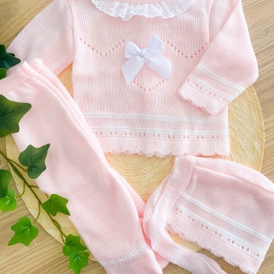 Conjunto rosa claro de roupa de bebé com gola branca rendada e laço, incluindo camisola, calças e touca