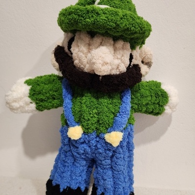 Boneco de pelúcia felpudo representando personagem com boné verde, camisa verde, calças azuis e sapatos pretos