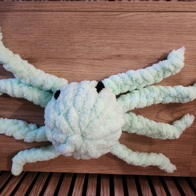 Brinquedo de pelúcia verde claro em forma de lula com olhos pretos sobre superfície de madeira.