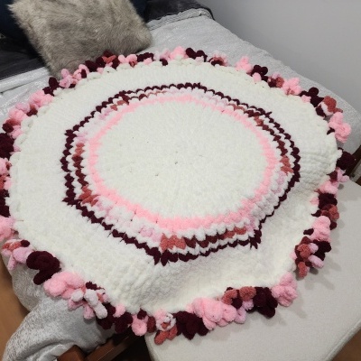 Tapete redondo de lã com pompons em cores branco, rosa e bordô numa cama cinza.
