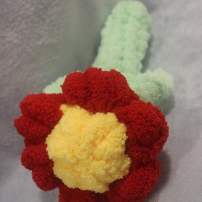 Brinquedo de pelúcia em forma de flor com pétalas vermelhas, centro amarelo e caule verde