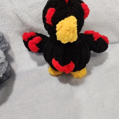 Pelúcia artesanal de pássaro preta com detalhes vermelhos e amarelos sobre tecido cinzento