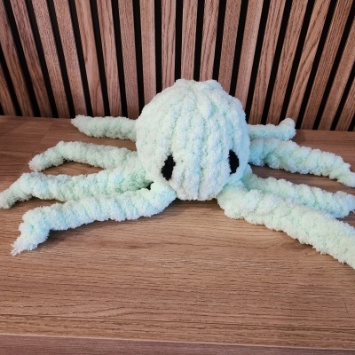 Pelúcia em formato de polvo felpudo branco com tons verdes e oito tentáculos sobre madeira