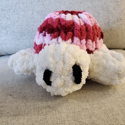 Pelúcia artesanal de tartaruga em lã branca, vermelha e rosa numa superfície cinza.
