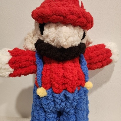 Boneco de malha tipo amigurumi vermelho, azul e branco contra parede.