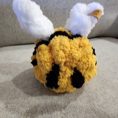 Boneco de pompons amarelos e pretos com asas brancas, em forma de abelha, sobre sofá cinzento.