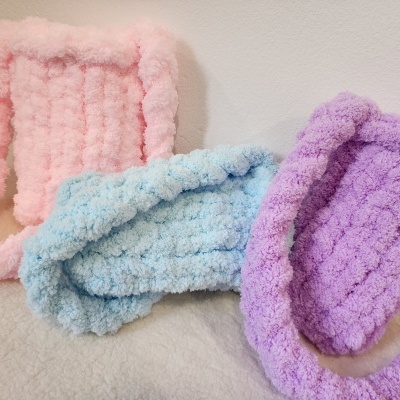 Três malas pequenas de peluche em rosa, azul claro e lilás sobre superfície creme.