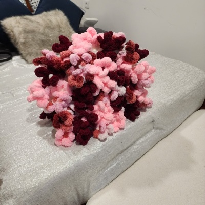 Almofada decorativa felpuda em tons de rosa e vermelho numa cama com roupa cinza prateada e almofadas azuis e bege