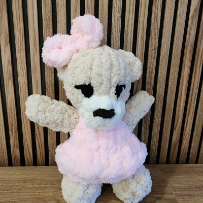 Urso de peluche castanho claro com vestido e laço rosa, olhos e nariz pretos